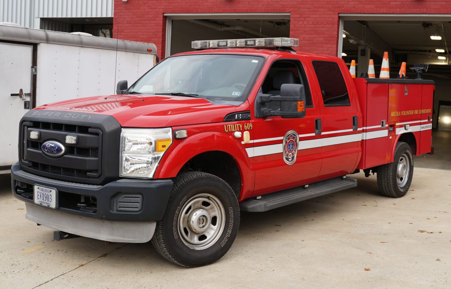 A red Ford Super Duty fire utility truck with emergency lights and orange traffic cones is parked outside a fire station with open garage doors.