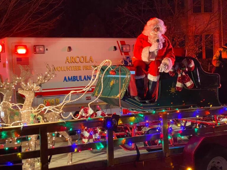 Santa Claus sits in a sleigh decorated with lights and candy canes on a float, with a lighted reindeer nearby and an ambulance in the background. Colorful holiday lights outline the scene.