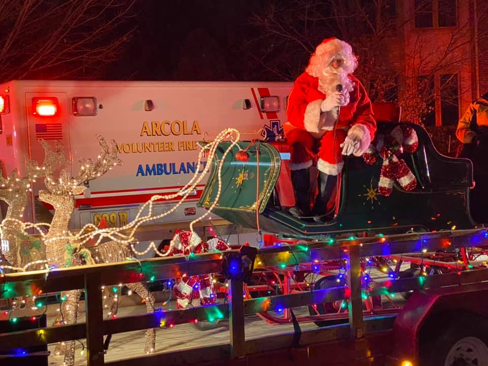 Santa Claus sits in a sleigh decorated with lights and candy canes on a float, with a lighted reindeer nearby and an ambulance in the background. Colorful holiday lights outline the scene.