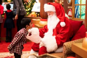 A young boy excitedly talks to Santa Claus, holding a letter, in a festive holiday setting decorated with Christmas trees and ornaments.