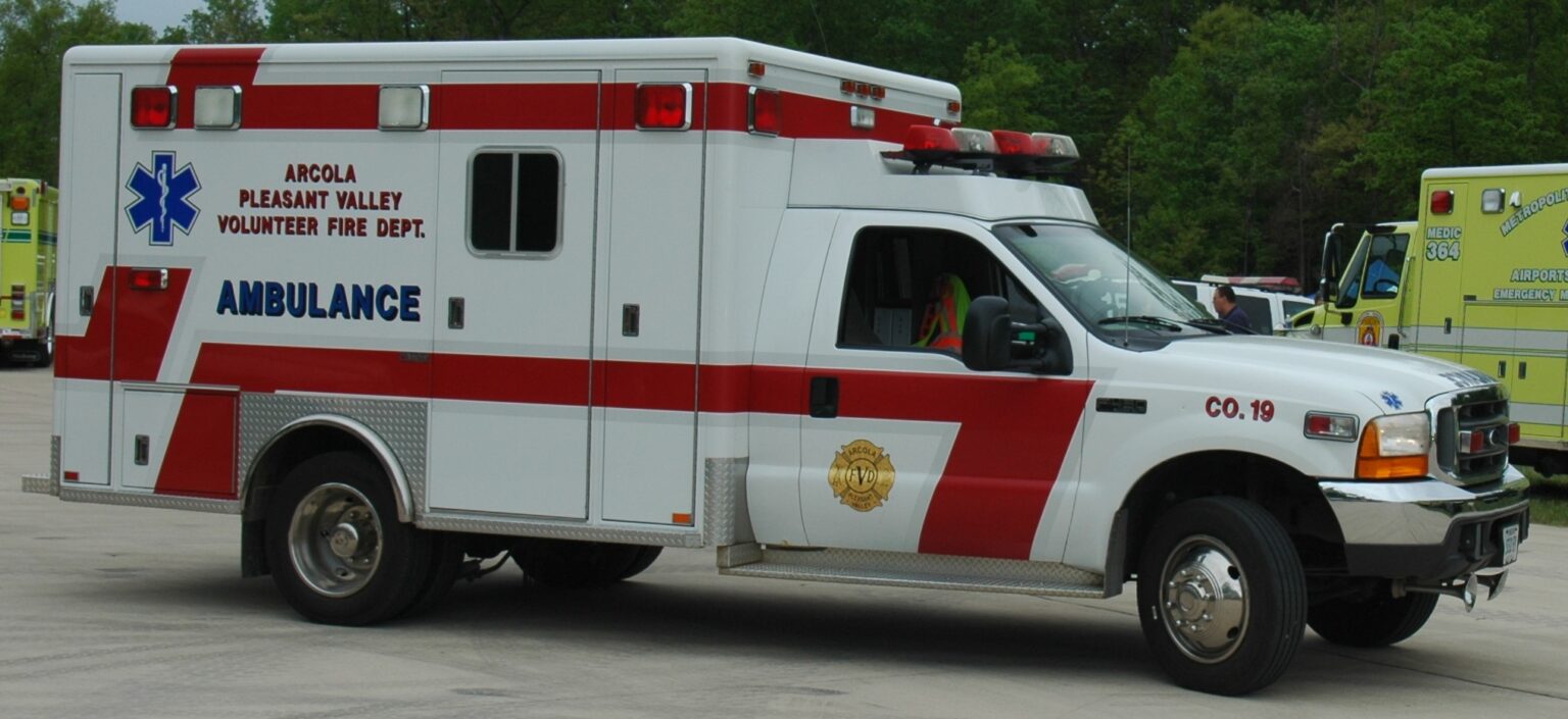 A white and red ambulance with “Arcola Pleasant Valley Volunteer Fire Dept.” on the side is parked on a paved surface. Other emergency vehicles are visible in the background.