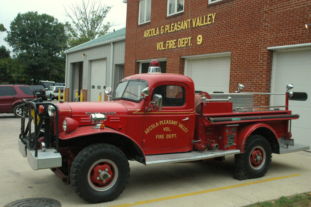 A vintage red Arcola-Pleasant Valley volunteer fire truck is parked outside a brick fire station with three garage doors and yellow signage.