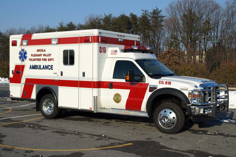 A white ambulance with red stripes and emergency lights is parked in a lot. The vehicle displays “Ambulance” and fire department markings on the side. Trees and snow are visible in the background.