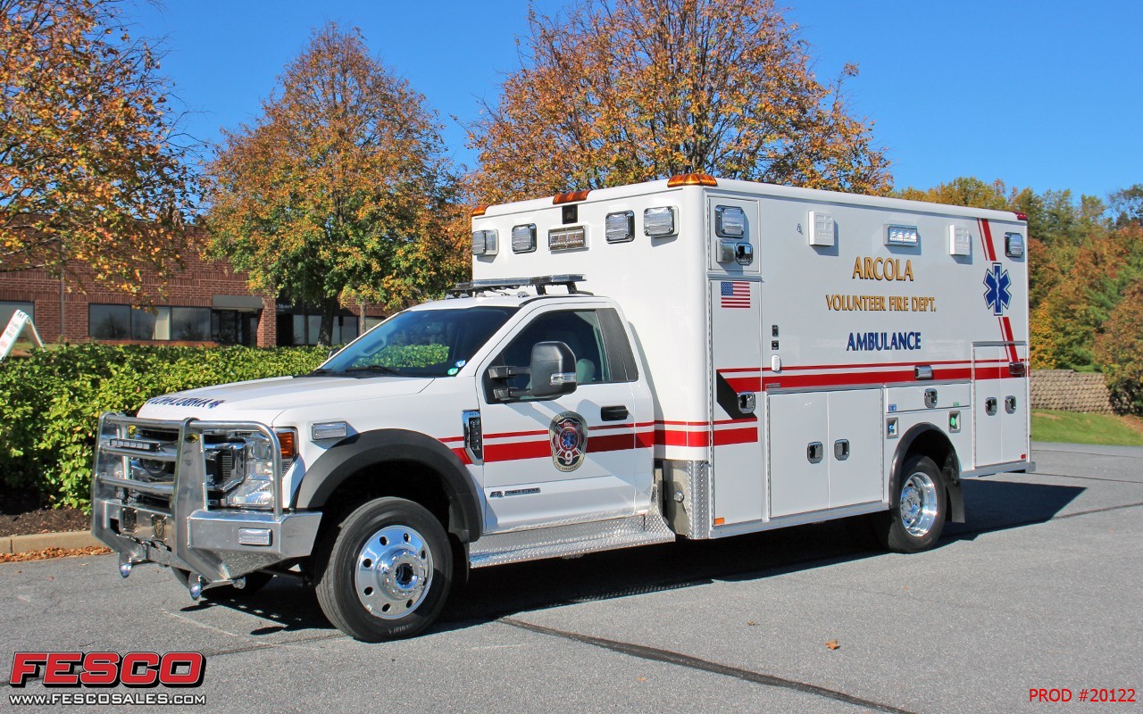 A white Alscola Volunteer Fire Dept. ambulance is parked on a street near trees with autumn leaves and a brick building in the background. The vehicle has red stripes and emergency markings.