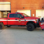 A red fire department truck with a snowplow attachment is parked outside a brick building. The truck has white stripes, emergency lights, and “MOOREA VOLUNTEER FIRE DEPARTMENT” written on its side.