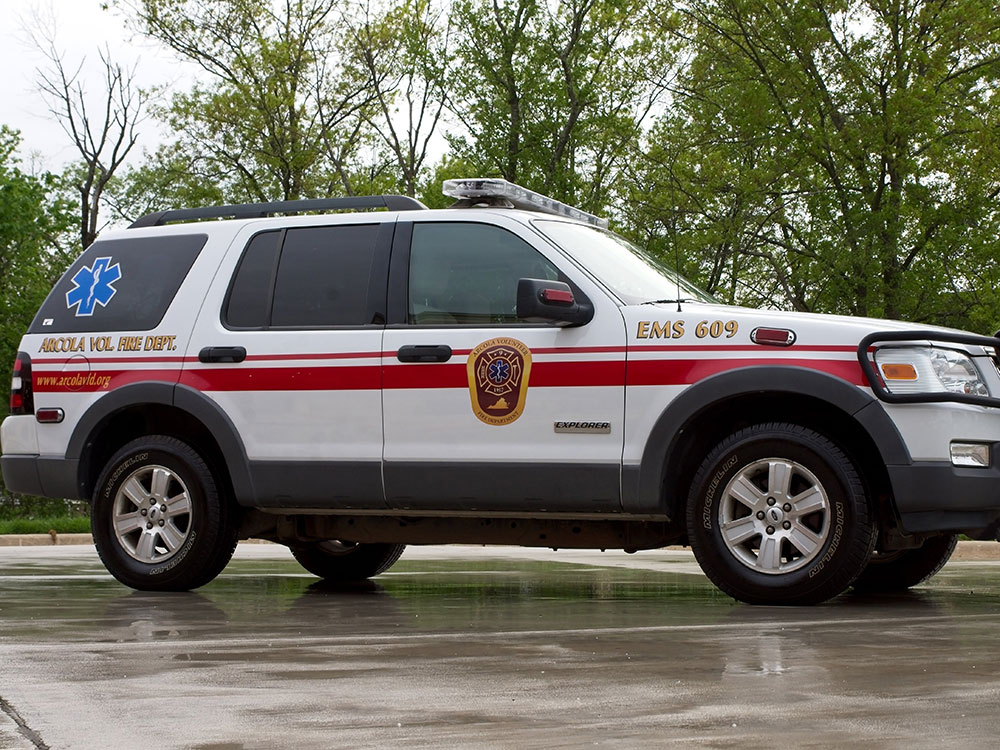 A white EMS SUV with red stripes and emergency symbols, parked on wet pavement near trees. The vehicle has “EMS 609” and “Azle Vol. Fire Dept.” written on it.