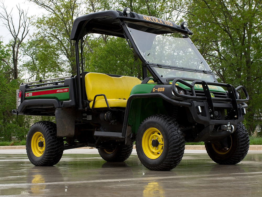A green and yellow utility vehicle with large tires and an open cab, parked on wet pavement with trees in the background.