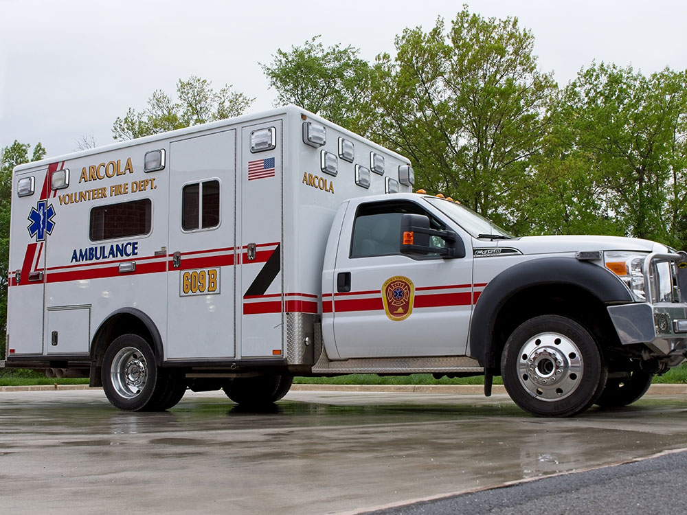 A white Arcola Volunteer Fire Dept. ambulance is parked on wet pavement, with trees and cloudy sky in the background.