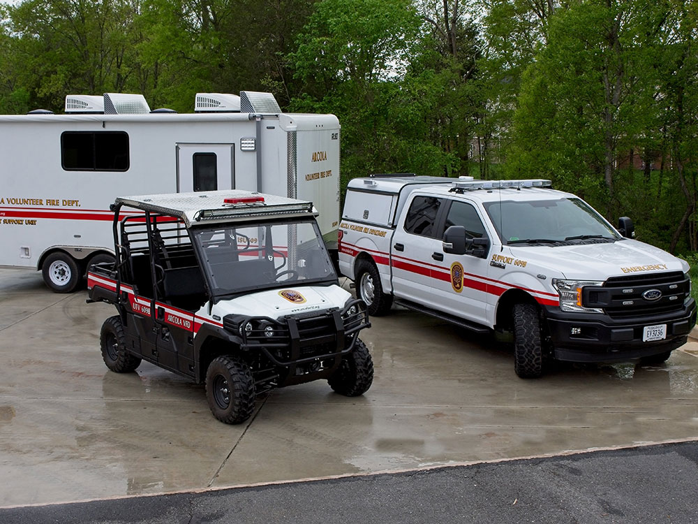 A volunteer fire department trailer, a white utility vehicle, and an ATV with fire department markings are parked on wet pavement near trees.