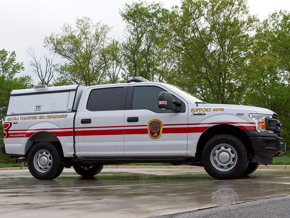 A white pickup truck labeled “Support 60-15” and “Volunteer Fire Department” is parked on wet pavement with trees and cloudy sky in the background.