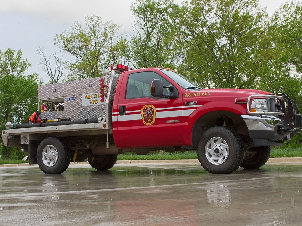 A red fire department brush truck with emergency lights is parked on wet pavement, surrounded by trees. The truck has equipment in the back and “Abcol VFD” is visible on the side.