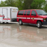 A red Arcola Volunteer Fire Department van is parked on wet pavement, towing a white trailer with department logos. Trees and greenery are visible in the background.
