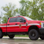 A red fire department pickup truck marked “Plow 609” is parked on a wet concrete surface, with trees and grass in the background.