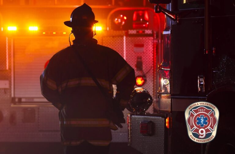 A firefighter in uniform stands near a fire truck with lights flashing at night. The badge of the Angola Volunteer Fire Department is visible in the lower right corner.