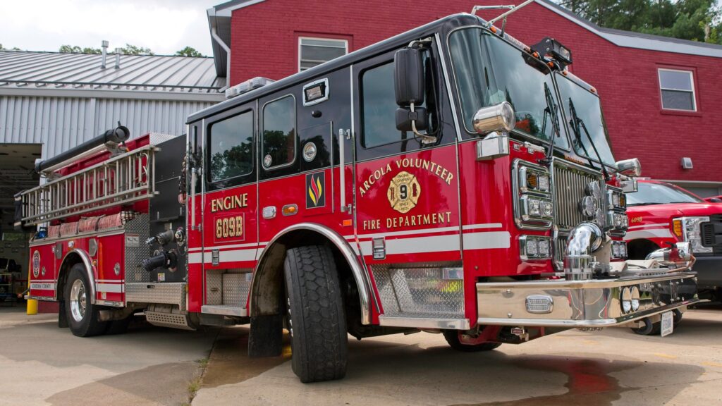 A red fire truck labeled “Tarcola Volunteer Fire Department” is parked outside a fire station with another emergency vehicle and building visible in the background.