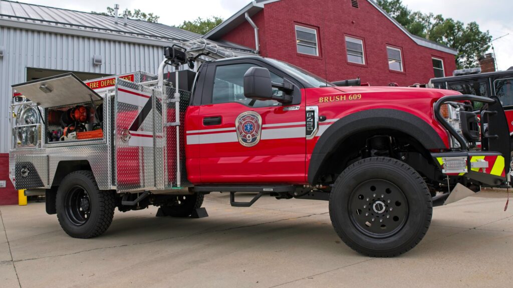 A red fire department brush truck with equipment compartments open is parked outside a brick fire station building. The truck has emergency lights, utility gear, and the fire department logo on its door.