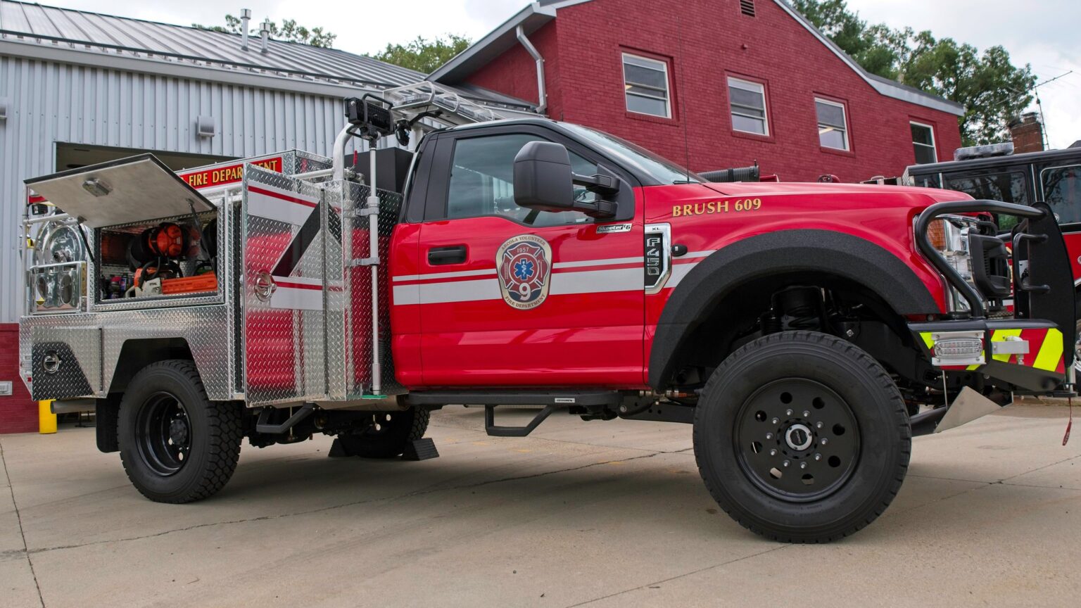 A red fire department brush truck with equipment compartments open is parked outside a brick fire station building. The truck has emergency lights, utility gear, and the fire department logo on its door.