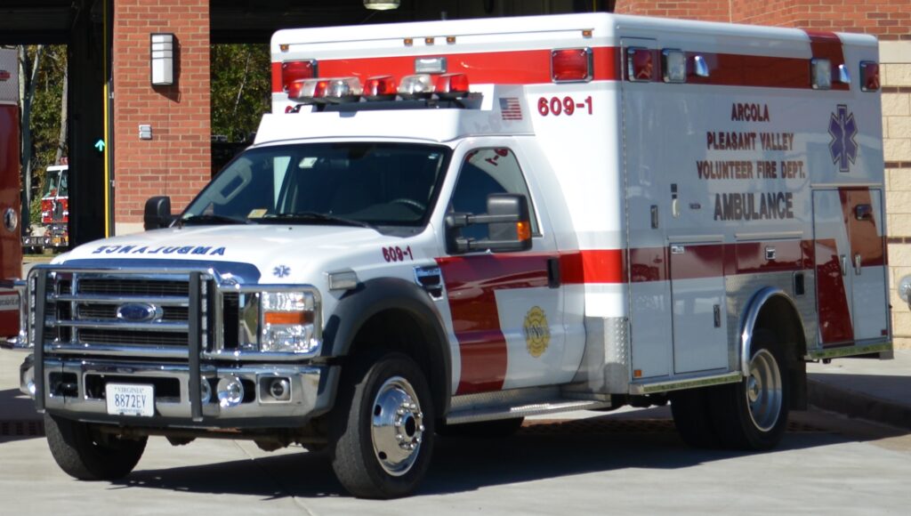 A white and red ambulance from the Arcola Volunteer Fire Department is parked outside a brick building, facing left. The vehicle has emergency lights on top and official markings on the side.