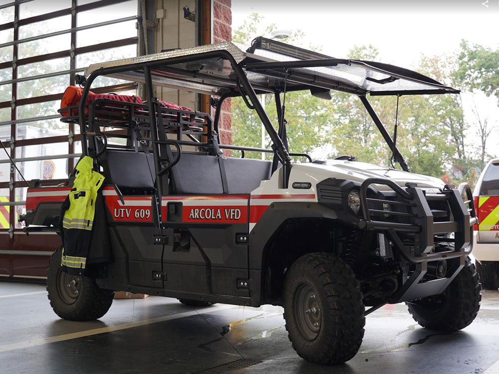 A utility vehicle labeled “UTV 609 Arcola VFD” with emergency lights and gear, including a yellow jacket, is parked inside a fire station near large windows.