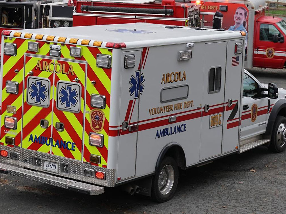 A white Arcola Volunteer Fire Department ambulance with red and yellow chevrons on the back is parked near a red fire truck. Emergency symbols and text are visible on the ambulance.