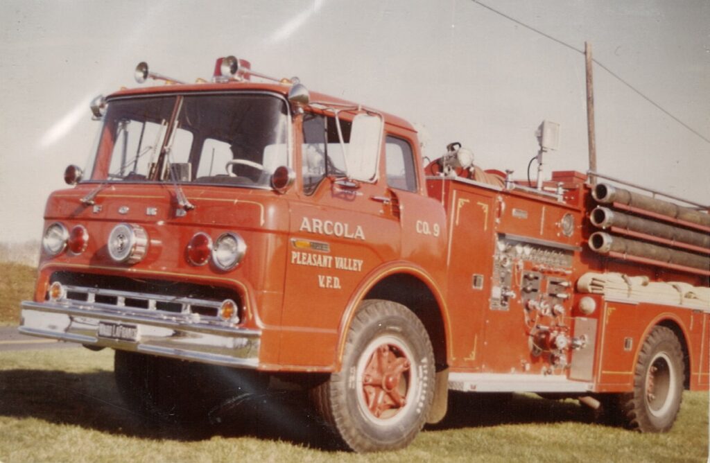 Vintage red fire truck labeled “Arcola Pleasant Valley V.F.D. Co. 9” parked on grass, with hoses and equipment mounted on the side.