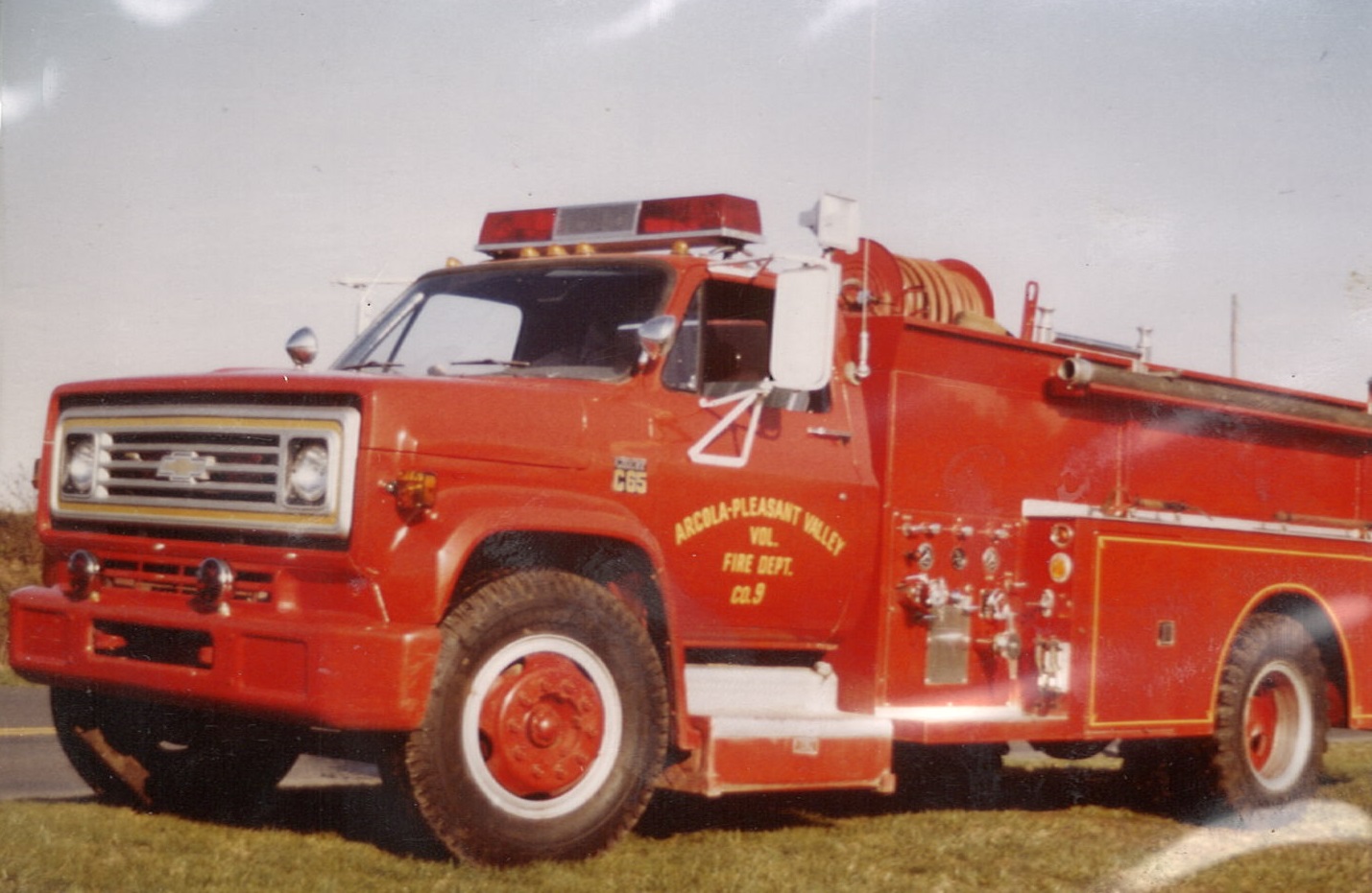 A vintage red fire truck with hoses and equipment is parked on a road. The truck has “AVOCA-PLEASANTVILLE FIRE DEPT.” written on its door and a visible siren on top.