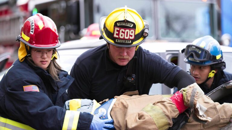 Three firefighters wearing helmets and gear assist a person on a stretcher near emergency vehicles, working together during a rescue operation.