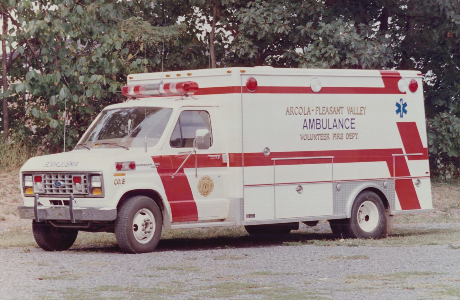 A white and red ambulance parked on a gravel surface, with “Arcola-Pleasant Valley Volunteer Fire Dept. Ambulance” written on the side. Trees and greenery are in the background.