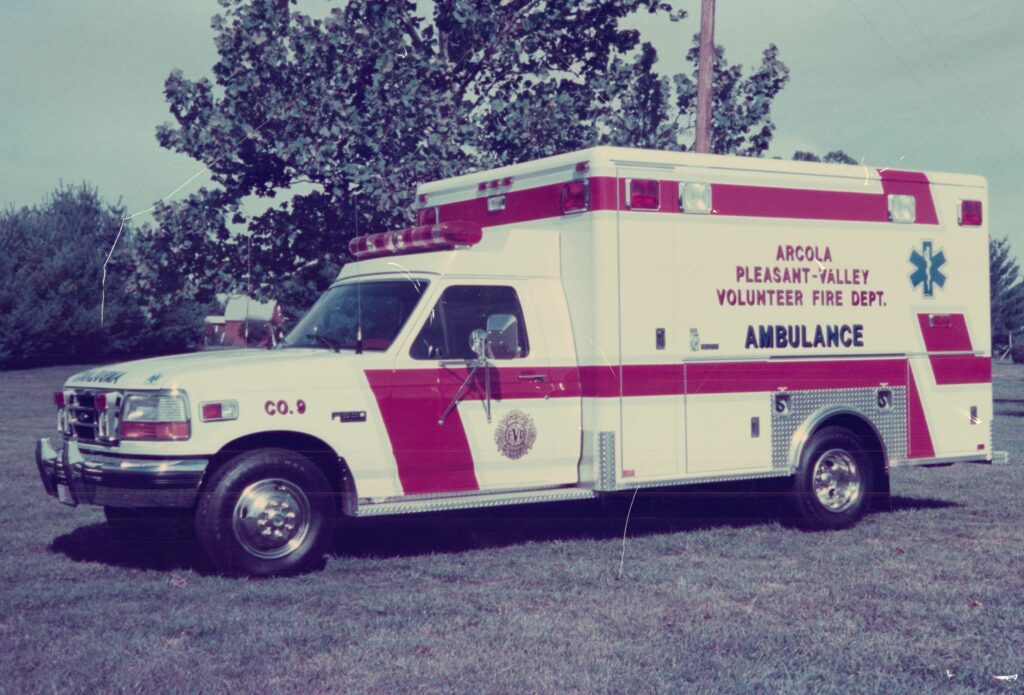 A white and red ambulance labeled “Arcola Pleasant-Valley Volunteer Fire Dept.” is parked on grass with trees in the background on a sunny day.