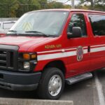A red van with white stripes and fire department markings is parked in a lot. The words “ADMIN 609” and department seals appear on the side. Trees and other vehicles are in the background.