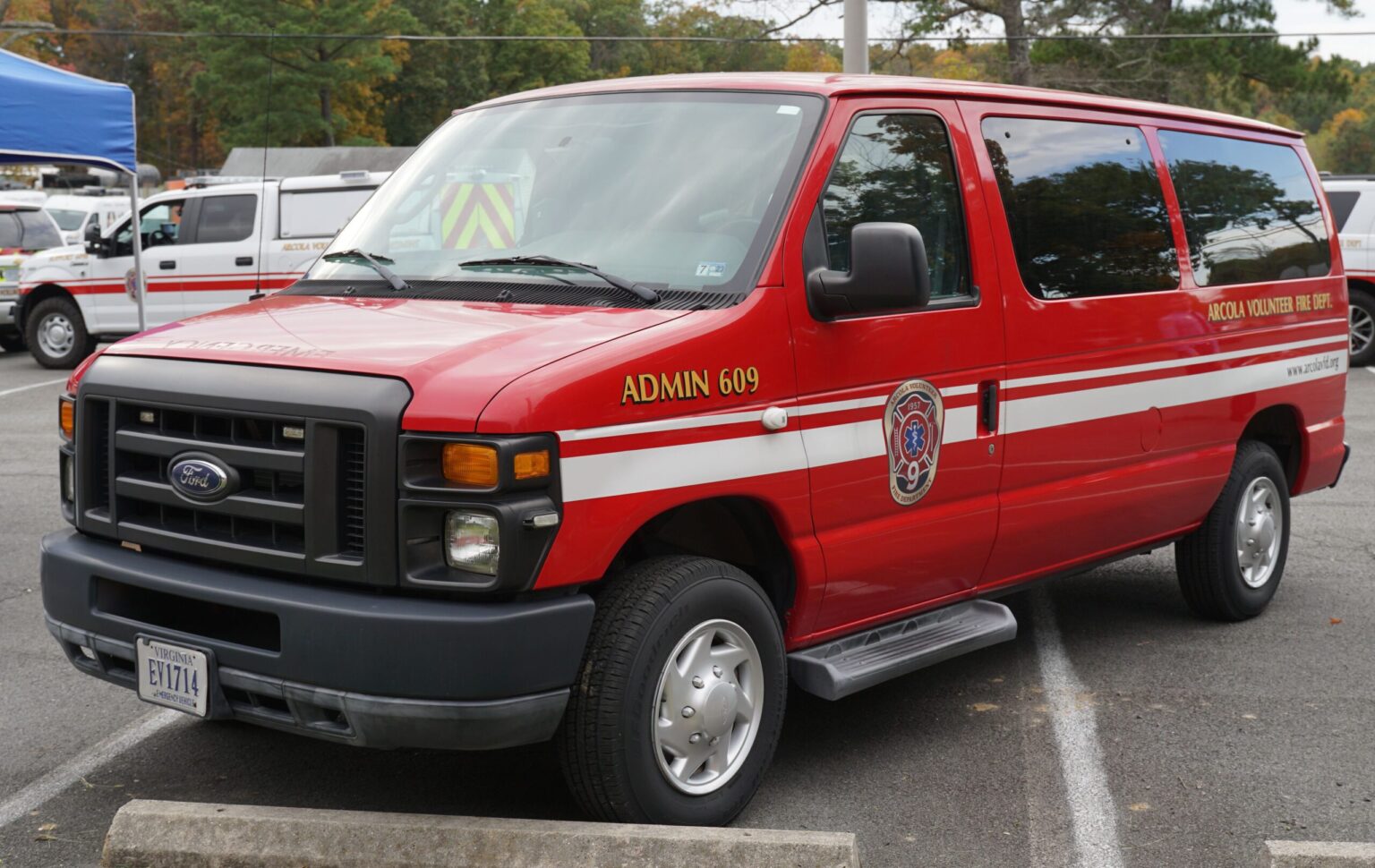 A red Ford van with white stripes, labeled “ADMIN 609” and “VOLUNTEER FIRE DEPT,” is parked in a lot. The van has official fire department logos and emergency lights on the roof.