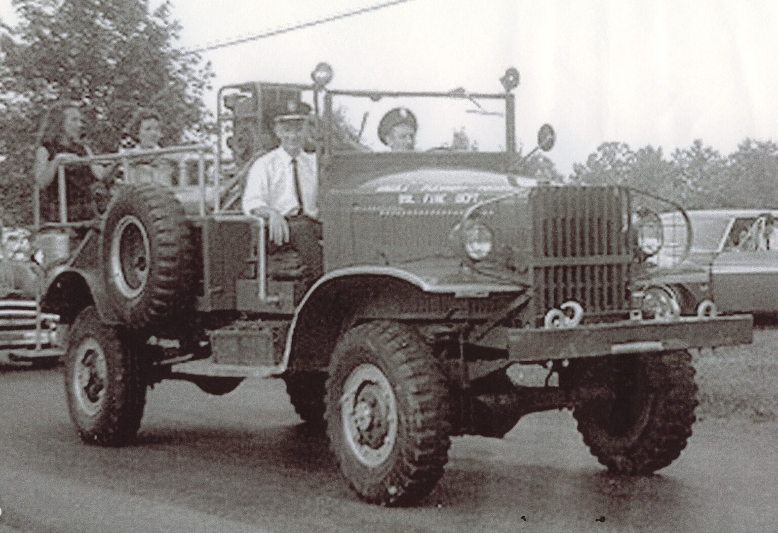 A vintage fire truck with several people riding on it is driving down a road. Trees and parked cars are visible in the background. The image appears to be from an earlier era, possibly mid-20th century.