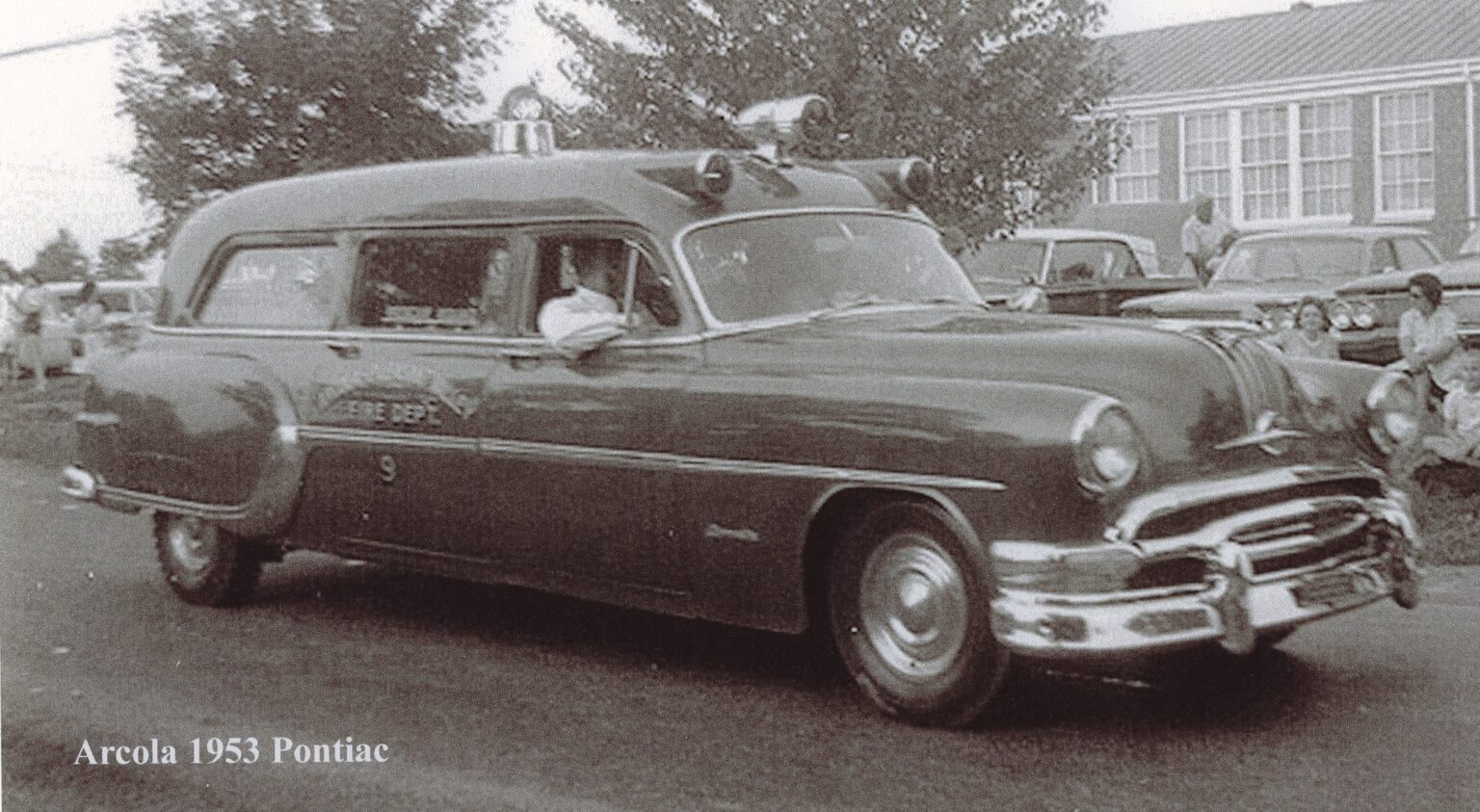 A vintage black-and-white photo shows a 1953 Pontiac ambulance labeled “FIRE DEPT” driving on a street, with a driver inside and classic cars and a building in the background.