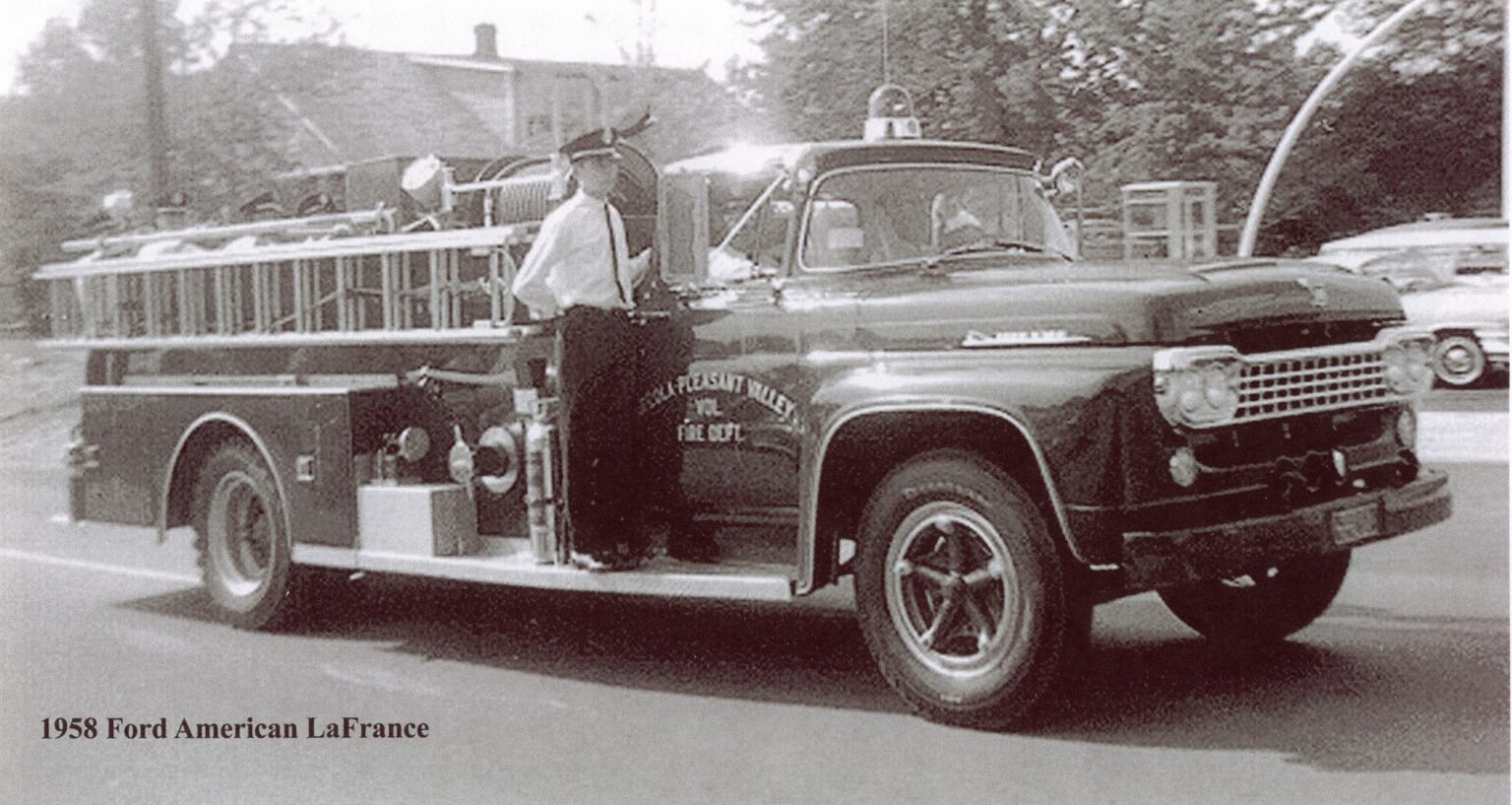 A vintage 1958 Ford American LaFrance fire truck drives down a street with a uniformed firefighter standing on the side. Trees and a building are visible in the background.