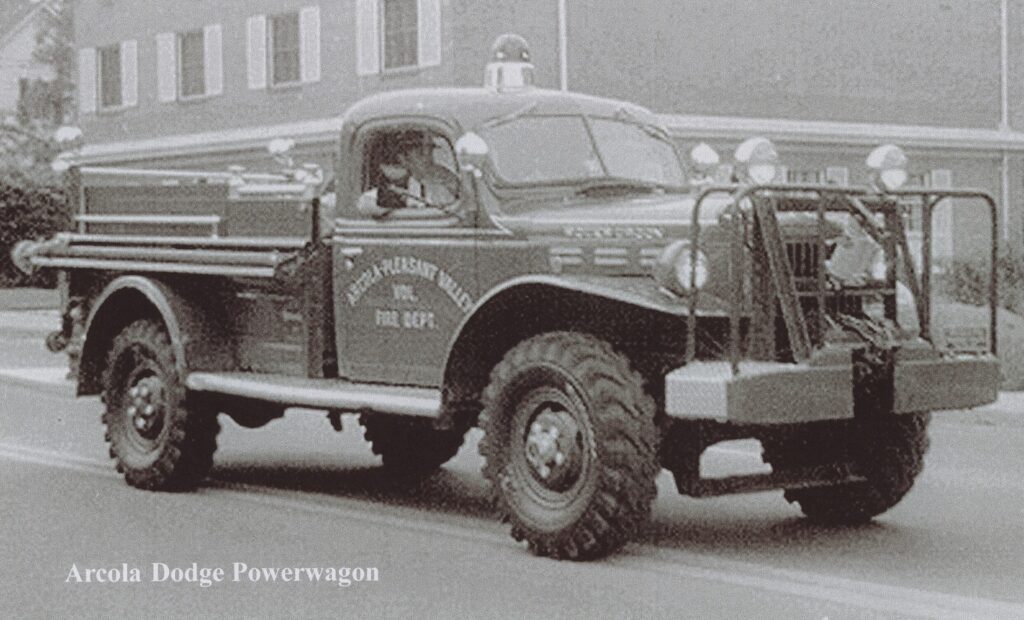 A vintage Dodge Power Wagon fire truck marked “Mt. Pleasant-Arcola Fire Dept.” drives down a street, equipped with large tires, front guards, and emergency lights.