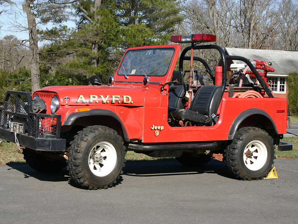 A vintage red Jeep with “A.P.V.F.D.” on the side, equipped with firefighting gear and hoses, parked on a paved surface with trees and a building in the background.