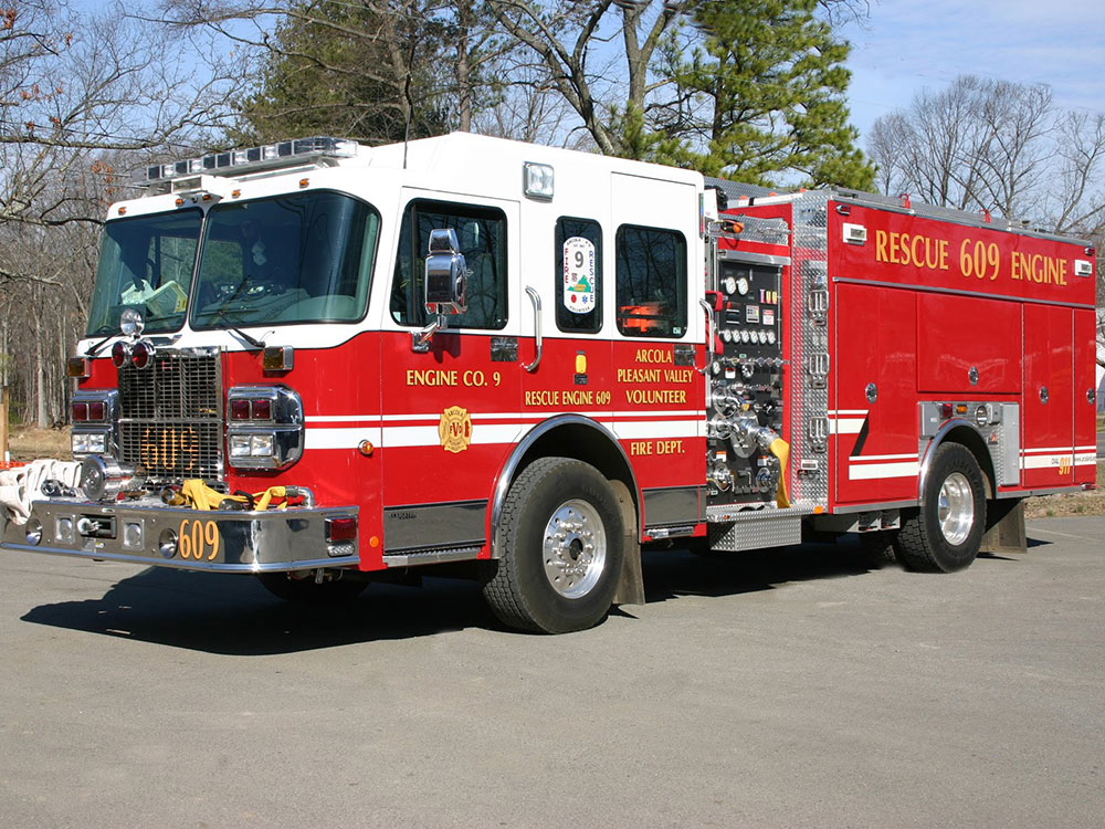 A large red fire engine labeled “Rescue 609 Engine” is parked outdoors on pavement, surrounded by trees and bushes. The vehicle displays emergency lights and reflective stripes, with various equipment compartments visible.