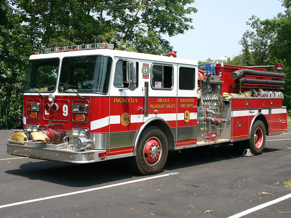 A red and white fire truck labeled “Engine Co. 9” parked on asphalt, with hoses and equipment visible, surrounded by green trees in the background.