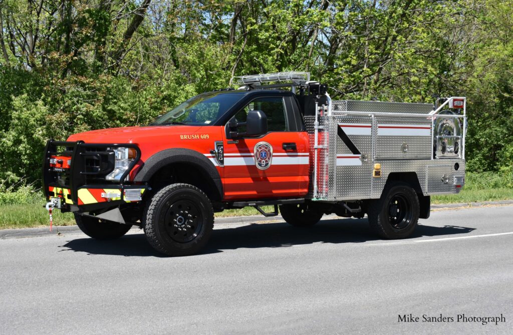 A red and silver brush fire truck is parked on a road with green trees in the background. The truck has emergency lights, a visible fire department emblem, and utility compartments on the side.