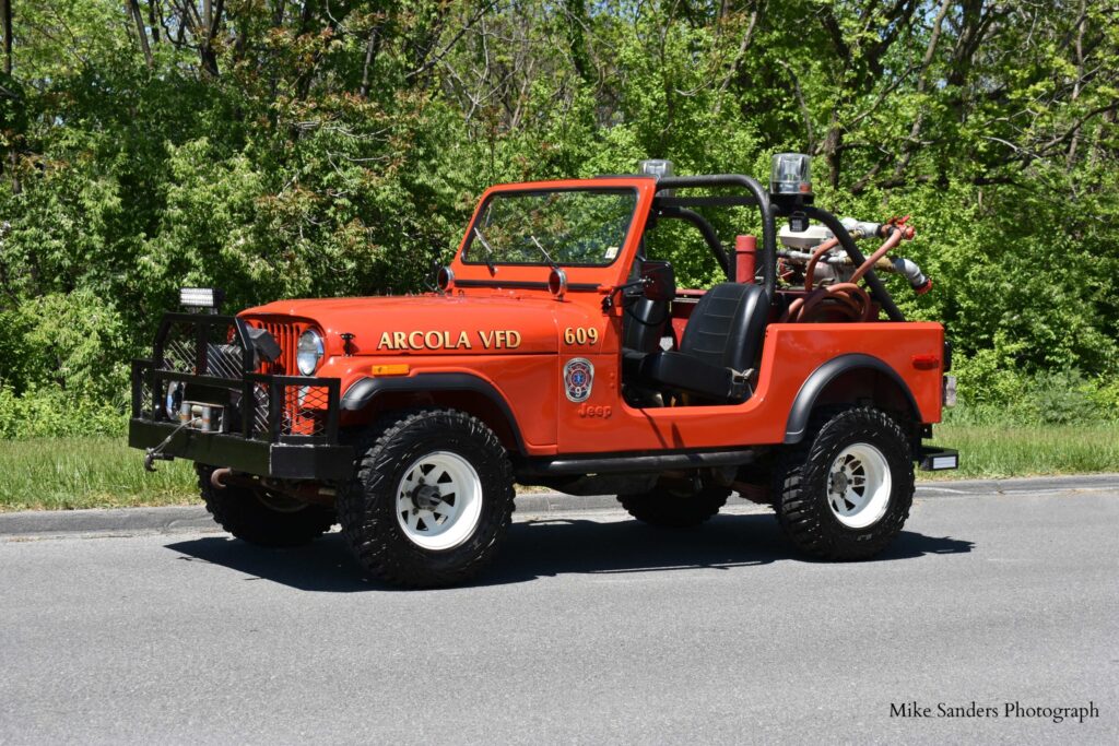 A red Arcola VFD fire Jeep with emergency equipment is parked on the roadside, surrounded by greenery and trees on a sunny day.