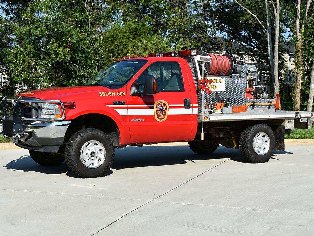 A red fire department brush truck with equipment and hoses on the back is parked on a paved surface. Trees and buildings are visible in the background.