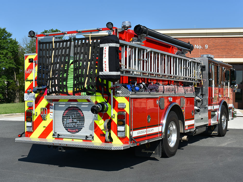 A red fire truck is parked outside a brick fire station. The truck has ladders, hoses, and equipment visible on the back, with reflective safety stripes and a number 9 on the building in the background.