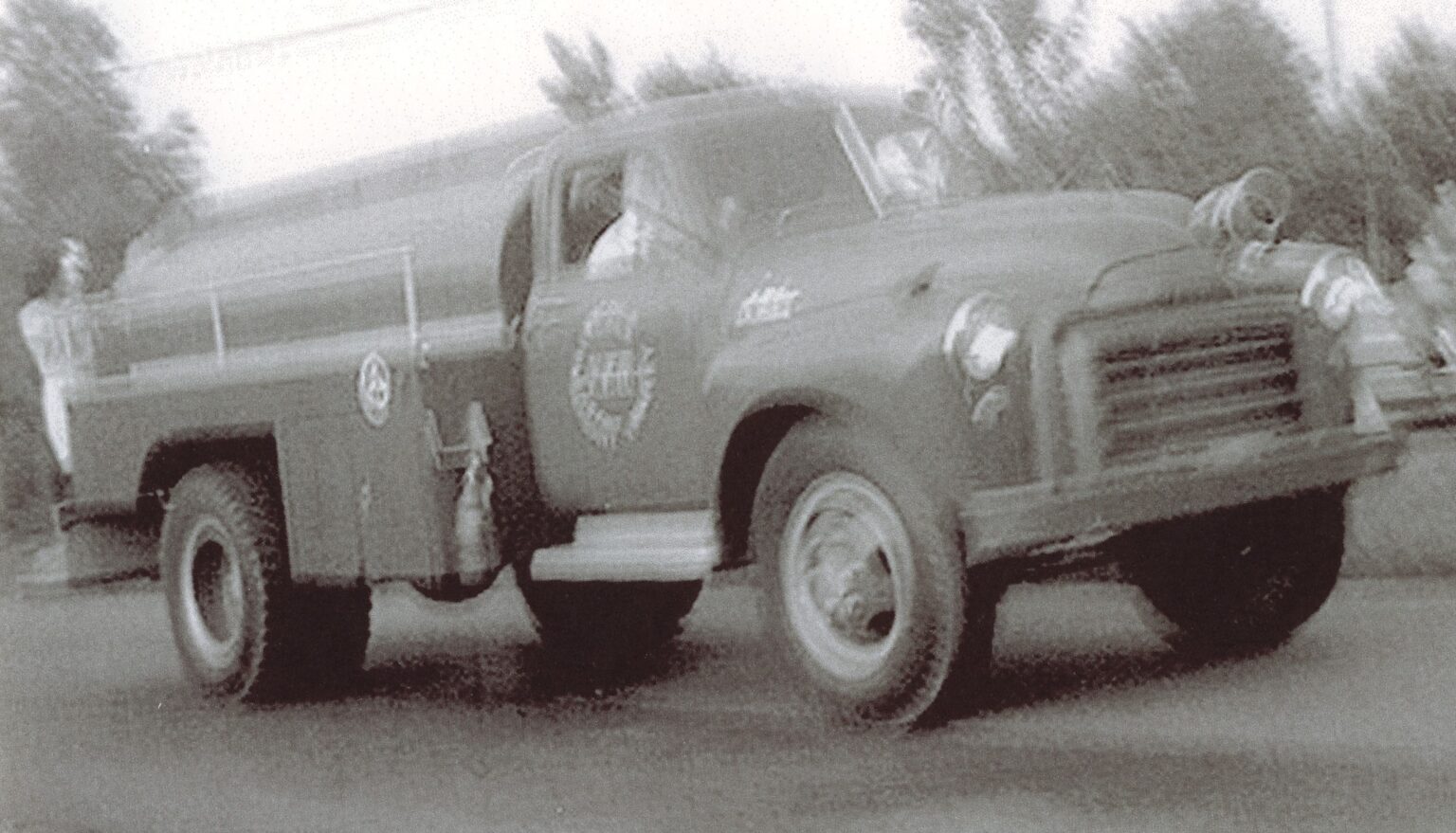 Black and white photo of a vintage fire truck in motion, with blurred background. The truck has a rounded front and emergency lights on top, and visible fire department logos on the door.
