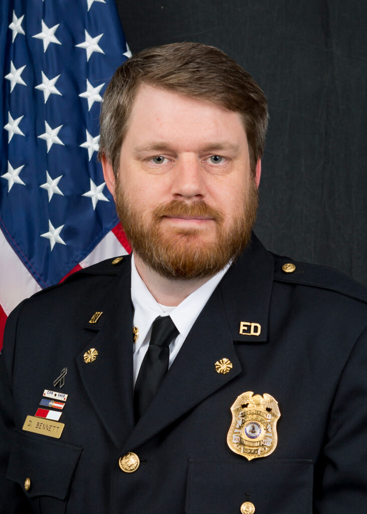 A man with brown hair and a beard, wearing a dark fire department dress uniform, stands in front of an American flag.