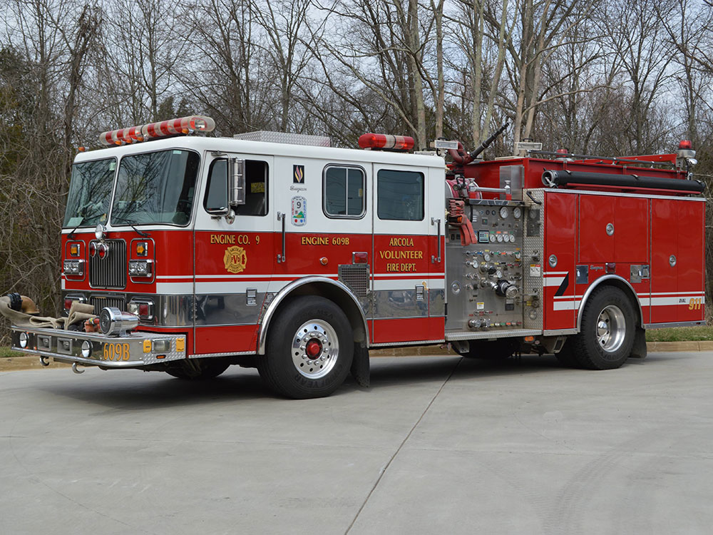 Red and white fire truck parked on a driveway with trees in the background. The truck belongs to a volunteer fire department and has hoses, dials, and emergency lights visible.