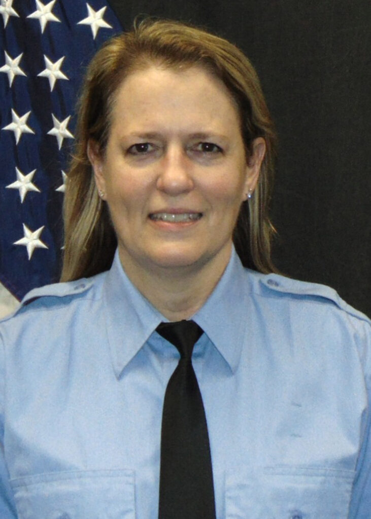 A woman in a light blue uniform shirt and black tie stands in front of a partially visible American flag, smiling slightly.