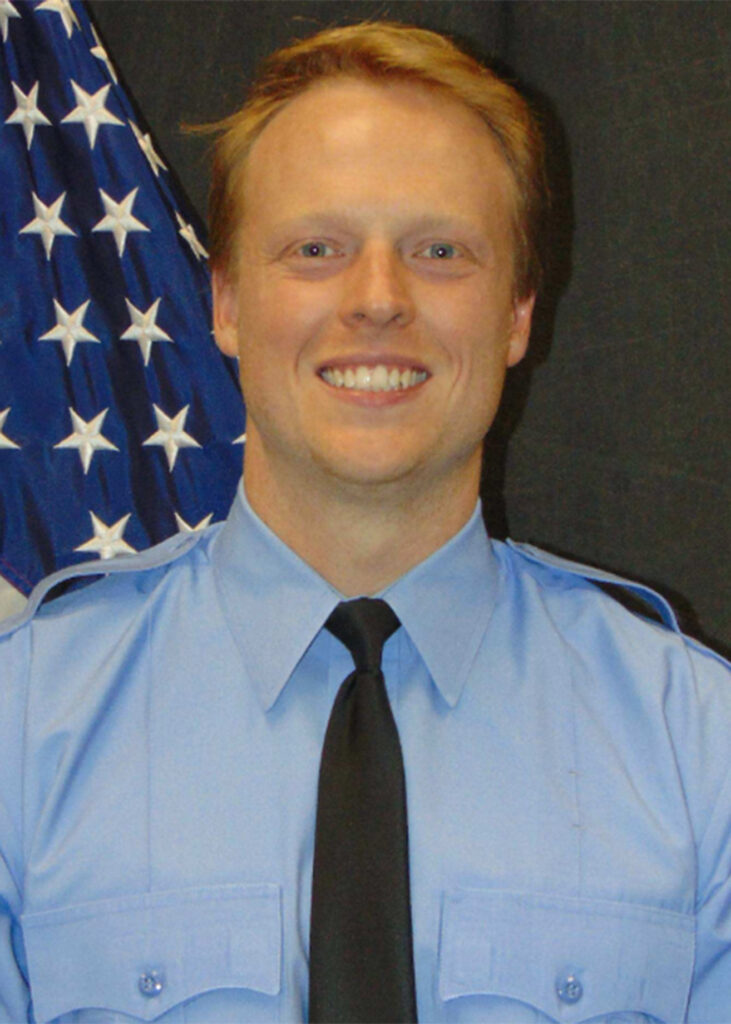 A smiling man in a light blue uniform shirt and black tie stands in front of a U.S. flag.
