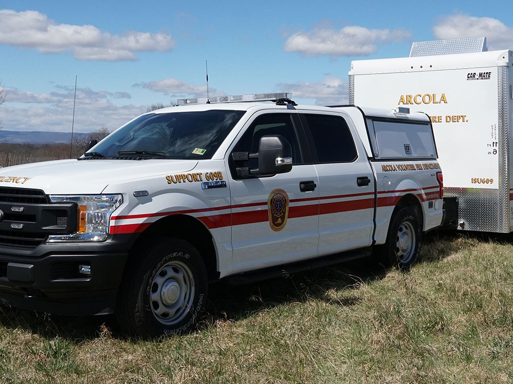 A white pickup truck with red stripes and fire department markings is parked on grass beside a white trailer labeled “ARCOLA VOLUNTEER FIRE DEPT.” under a partly cloudy sky.