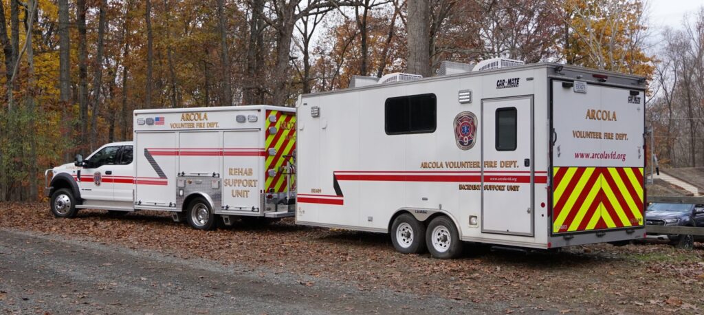 Two Arcola Volunteer Fire Department emergency response vehicles, including a rehab/support unit and a command unit, are parked on a leaf-covered ground in a wooded area during autumn.
