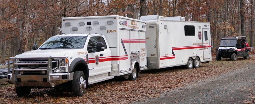 A large white emergency response vehicle and trailer with red stripes are parked on a leaf-covered path in a wooded area; another off-road vehicle is visible behind them.